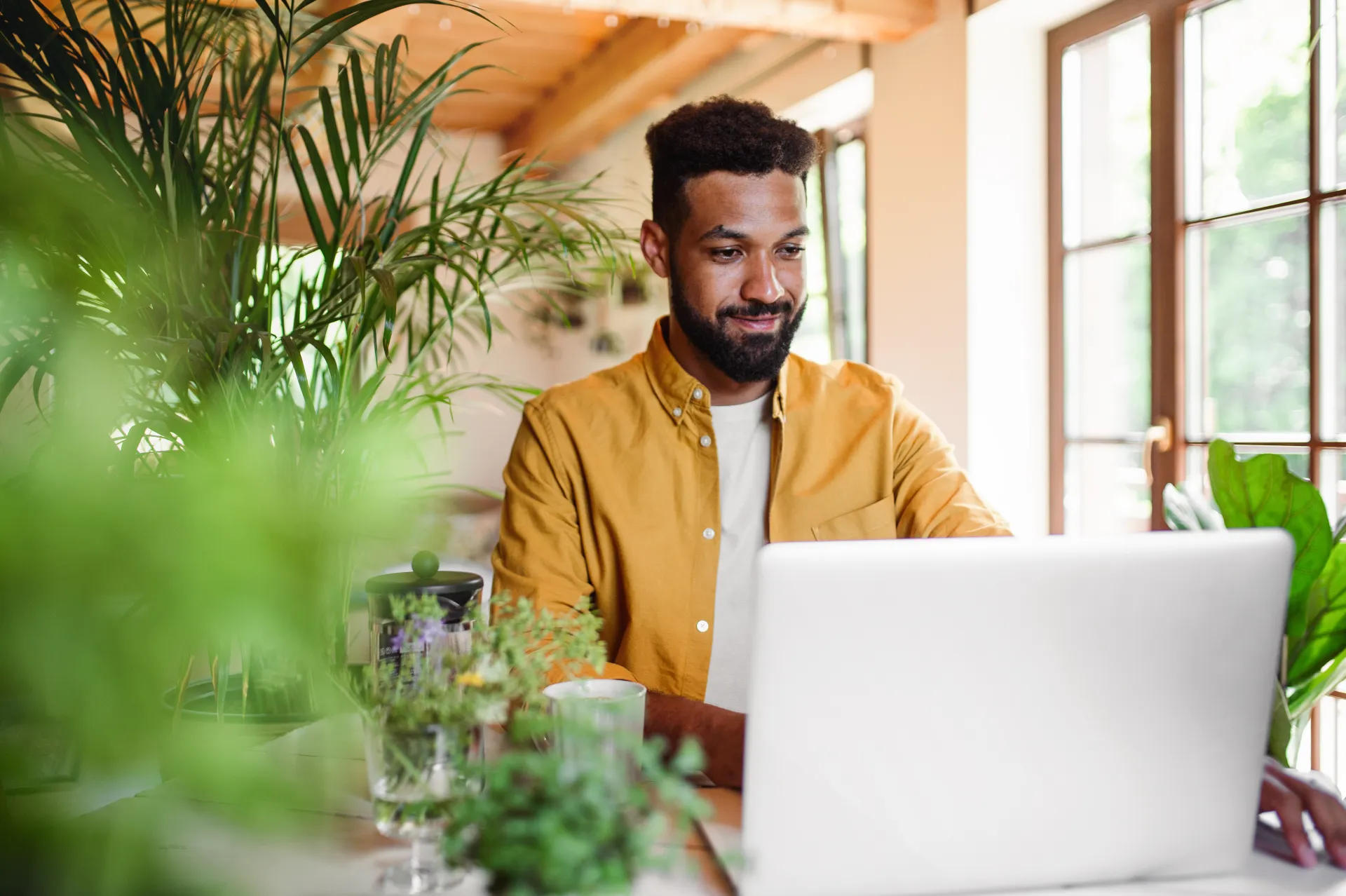 A young black man wearing a yellow button-up shirt over a white t-shirt sits at a table in a room with bright natural light surrounded by indoor plants working on his laptop.