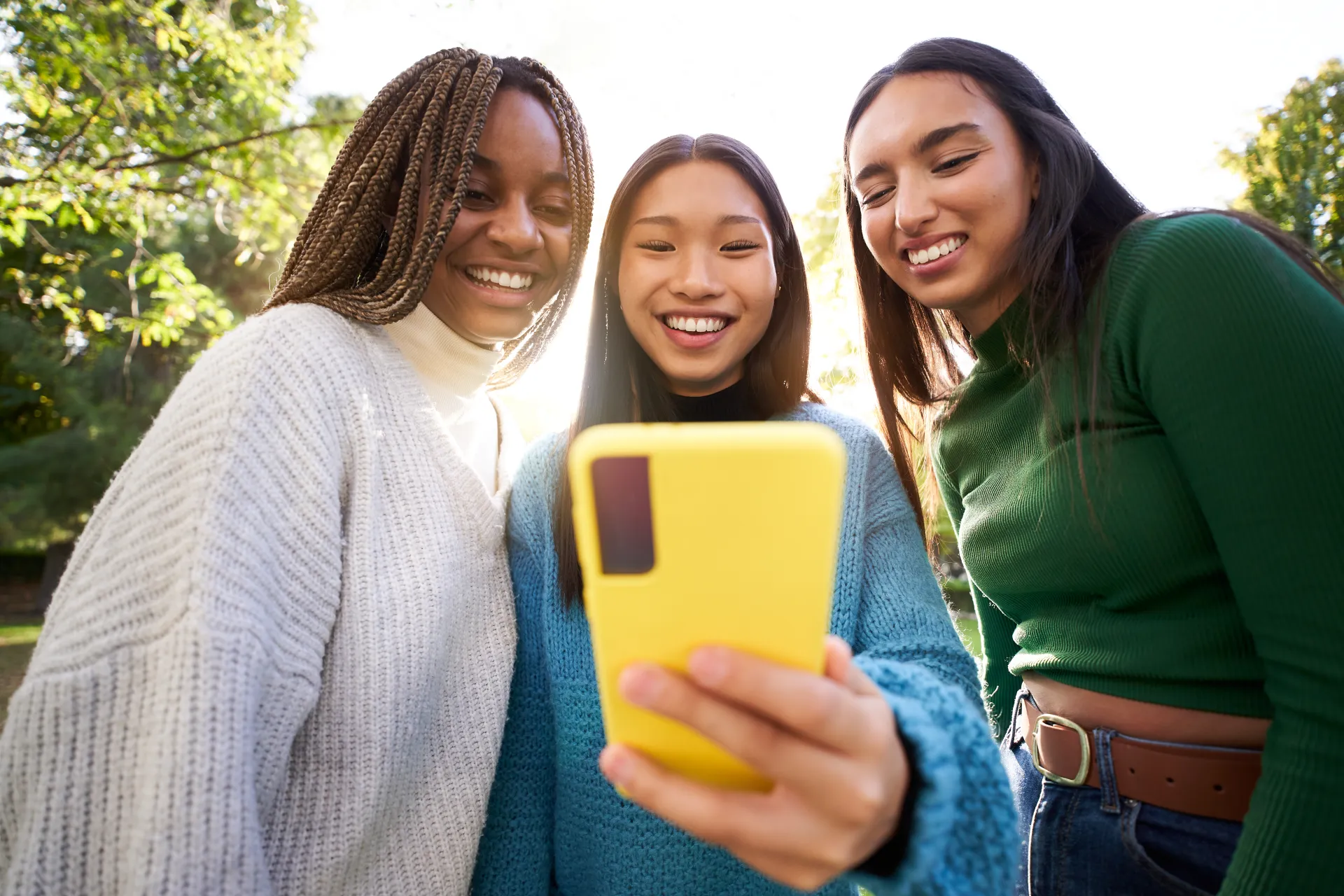 A young Asian woman in a blue sweater holds out a yellow smartphone while a black woman in a white sweater and a woman with dark hair in a green shirt look at the screen.