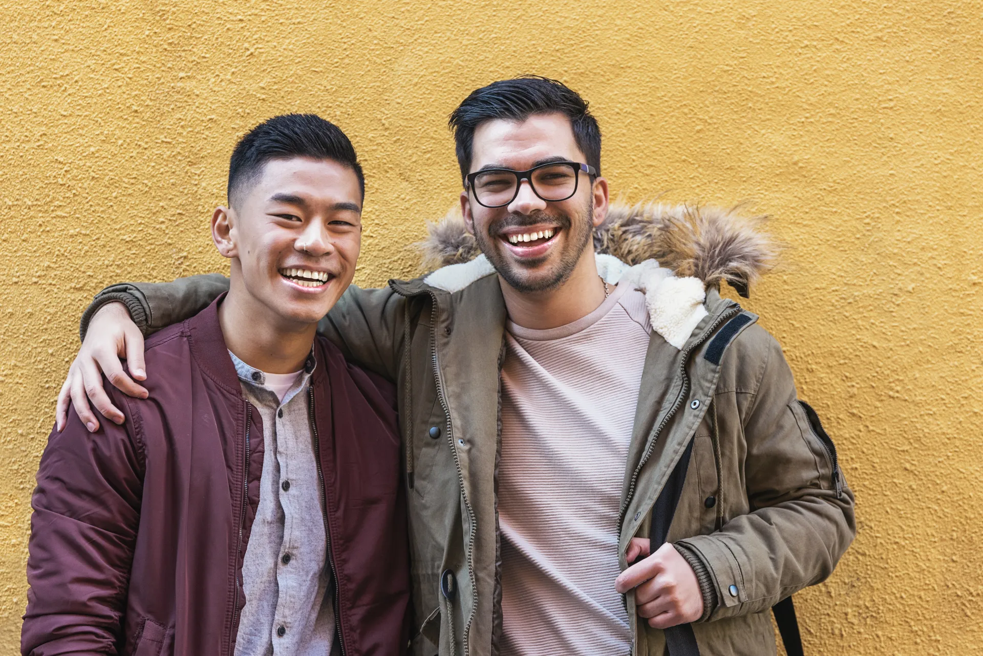Two young men stand wearing coats laugh while standing with their arms around each other against a yellow stucco wall.